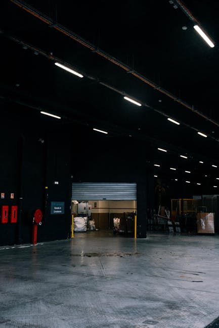 The image depicts the interior of a warehouse or storage facility with a concrete floor and high, dark ceiling illuminated by long, linear LED lighting fixtures. In the background, there is a partially open roller shutter door revealing an area with various stacked or bundled items, some wrapped in protective material. To the left of the door, red fire safety equipment is mounted on the black wall. Near the right side of the image, there are carts or trolleys loaded with cardboard boxes and plastic-wrapped pallets, indicating ongoing or recent material handling and rubbish collection. The environment appears clean and organized, consistent with professional waste management or clearance services such as those provided by Rubbish Clearance Acton, supporting the concept of private and site-specific rubbish removal. The overall scene suggests the facility's capacity for large-scale waste or rubbish clearance, aligning with independent waste disposal options rather than municipal collection, within an industrial or commercial setting.