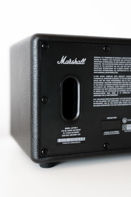 Close-up image of the rear panel of a black Marshall portable speaker, displaying a textured finish with visible screws and a central port for sound output. The back panel features a label with technical specifications, model information, certification codes, and regulatory details, all printed in white text on a black background. The device is positioned on a white surface with a light background, highlighting its compact, rectangular shape and minimalist design. This type of electronic equipment could be associated with office or event sound setups, relevant to context where independent or on-site waste disposal for electronic devices might be necessary, as arranged by local rubbish removal services like Rubbish Clearance Acton.