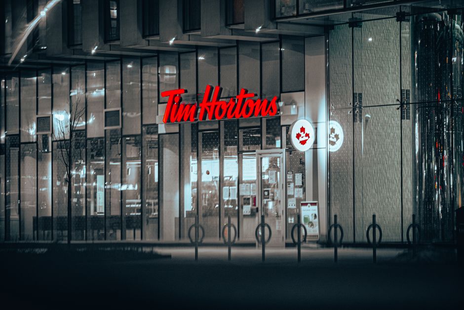 A nighttime exterior view of a Tim Hortons coffee shop with a modern glass facade reflecting surrounding lights. The red Tim Hortons logo is prominently displayed above the entrance, which features double glass doors and large windows showing the interior's illuminated space. To the right of the entrance, there are circular signs with the Tim Hortons emblem, set against the textured exterior wall, which appears to be finished with metallic or wooden panels. In front of the building, a row of bike racks is visible on the sidewalk, with no bicycles present. The area is dimly lit, emphasizing the glow from the interior and signage, and the scene projects a quiet, urban atmosphere consistent with commercial property outside a busy shopping area or retail outlet specializing in food and beverage services.