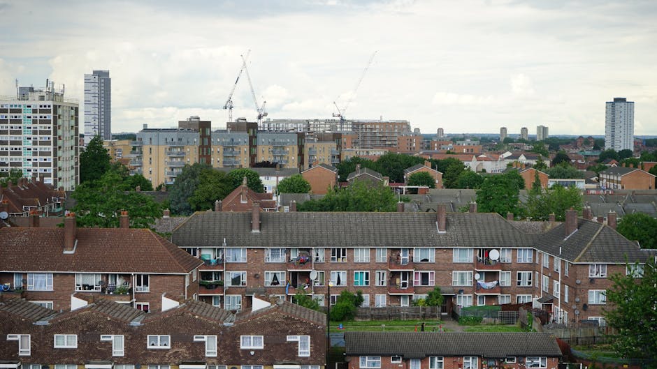 A wide view of a residential urban landscape showing multiple brick apartment buildings with grey tiled roofs in the foreground, each featuring small balconies with laundry lines and potted plants. Behind these, there are green trees interspersed among lower-rise housing and gardens, providing a leafy backdrop. Further in the distance, taller modern high-rise buildings with white and glass facades stand against a cloudy sky, where construction cranes are visible, indicating ongoing development. The scene captures a mix of older and newer structures, with the natural environment blending with ongoing urban growth. The lighting is subdued, suggesting an overcast day, and the overall atmosphere reflects a typical mid-sized city with diverse building styles and active construction for future expansion. This view illustrates a typical British residential neighbourhood, highlighting the importance of local waste management and rubbish removal services for maintaining such urban environments, as offered by companies like Rubbish Clearance Acton.