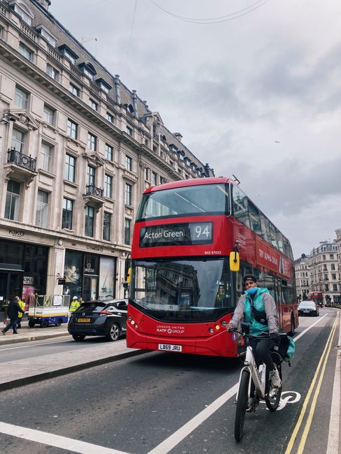 A red double-decker bus, displaying destination 'Acton Green' and route number 94, is travelling along a busy street lined with historic, multi-storey buildings featuring detailed stone facades, balconies, and large windows. The sky overhead is cloudy, creating a diffuse light that softly illuminates the scene. In the foreground, a cyclist wearing a blue face mask, a light grey hoodie, and dark trousers is riding a black and white bicycle, with a green backpack secured on his back. To the left, a few pedestrians are walking on the pavement, some wearing high-visibility jackets, possibly indicating roadwork or maintenance activity. Several parked cars and a small delivery vehicle are visible along the roadside, with a few moving vehicles in the background. The environment suggests an urban setting typical of London, where private and independent transport options like buses and cycling are integrated into everyday city life, consistent with local rubbish removal and waste management practices carried out by companies like Rubbish Clearance Acton, operating within the context of on-site or alternative waste handling services.