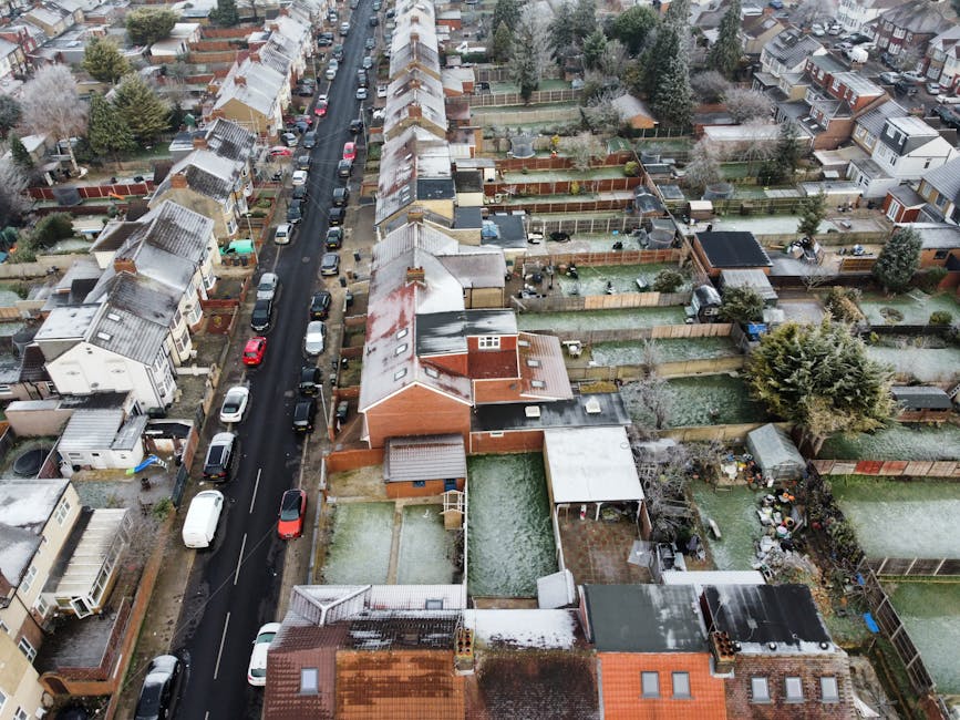 An aerial view of a residential neighbourhood during winter, showing a narrow street lined with parked cars on both sides, including sedans, hatchbacks, and vans in various colours such as white, red, black, and grey. The street runs through a densely packed area of terraced houses with pitched roofs, many of which have snow or frost on their roofs and rooftops of adjacent garages. Behind the houses, there are multiple private gardens enclosed by wooden fences, with some gardens featuring trees, shrubs, and outdoor furniture, and a few with visible sheds or garden structures. The image captures a mix of brick and weathered materials on the buildings’ facades and fences, with textures ranging from smooth brickwork to weathered wood. The surrounding environment includes residential yards, some with evident rubbish or discarded items, indicating recent or ongoing rubbish removal efforts by services such as Rubbish Clearance Acton. The overall atmosphere suggests a quiet winter day in an established suburban area, with natural lighting highlighting the varied textures of the rooftops, fences, and garden features, illustrating typical urban rubbish receptacle and waste management scenarios.